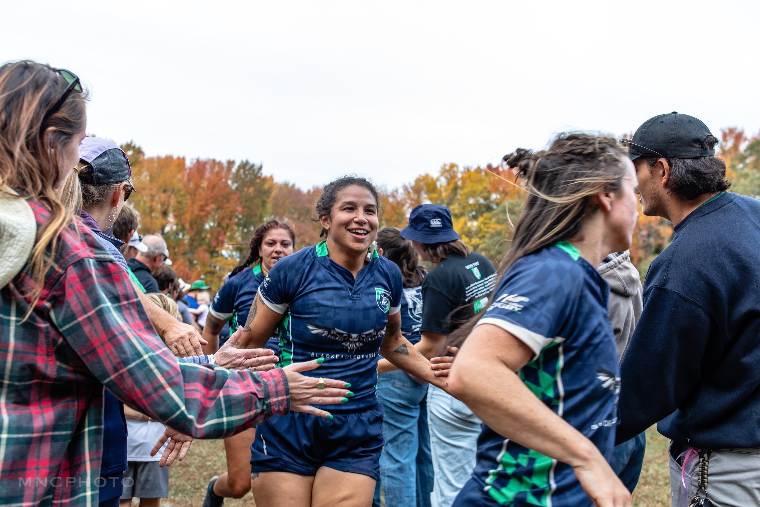 Women rugby players being congratulated by their family, friends, fans, and coaches after a rugby match with fall foliage in the background.