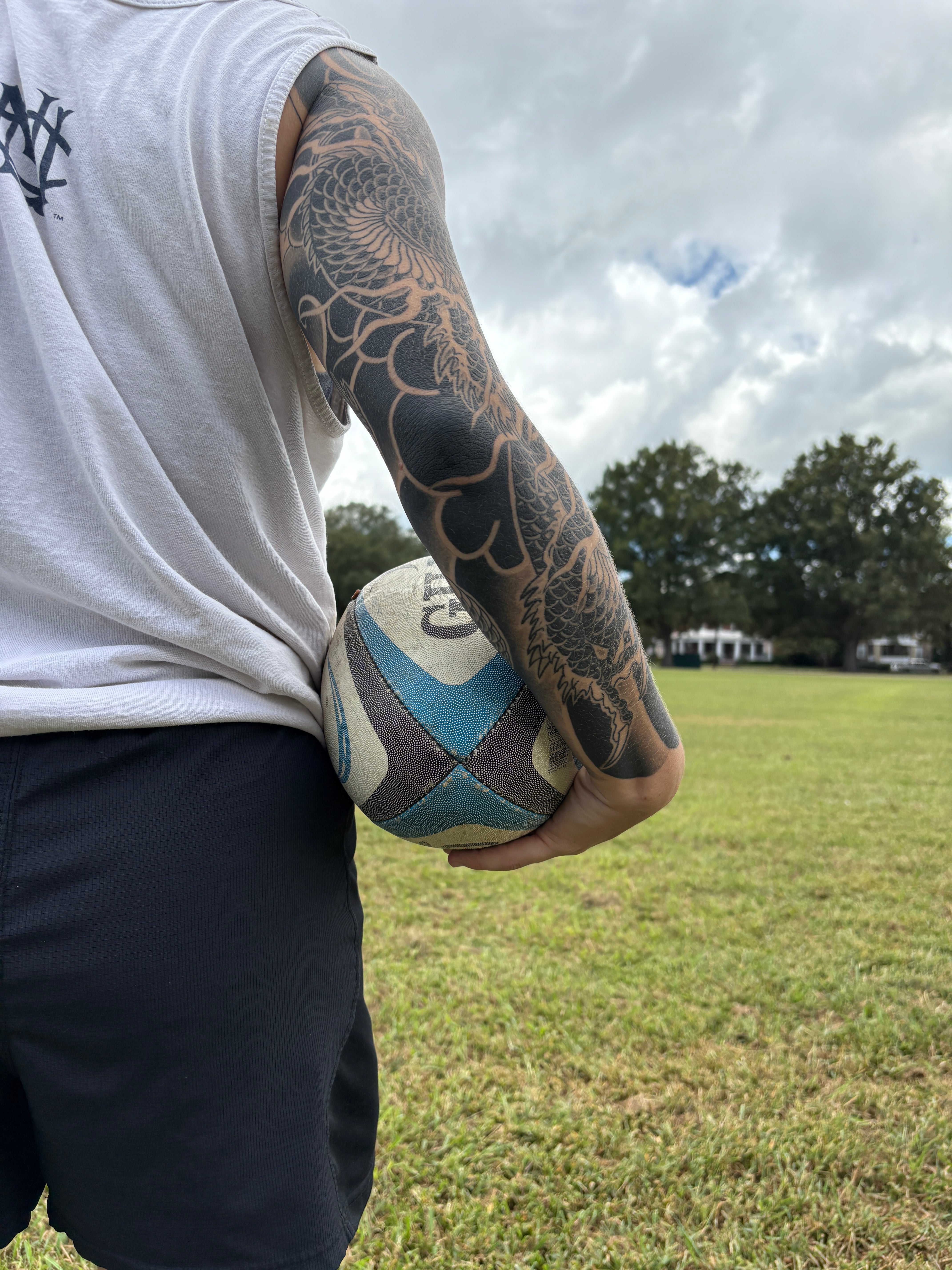Tattooed arm holding a rugby ball in a field outdoors.