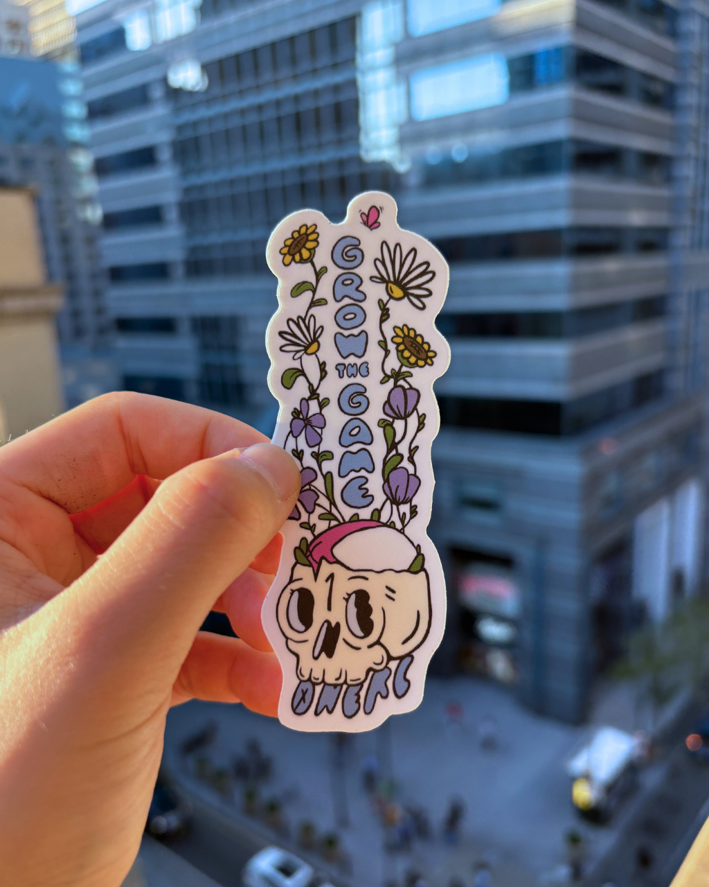 Hand holding a sticker of wildflowers growing out of a skull with a rugby ball resting in it.