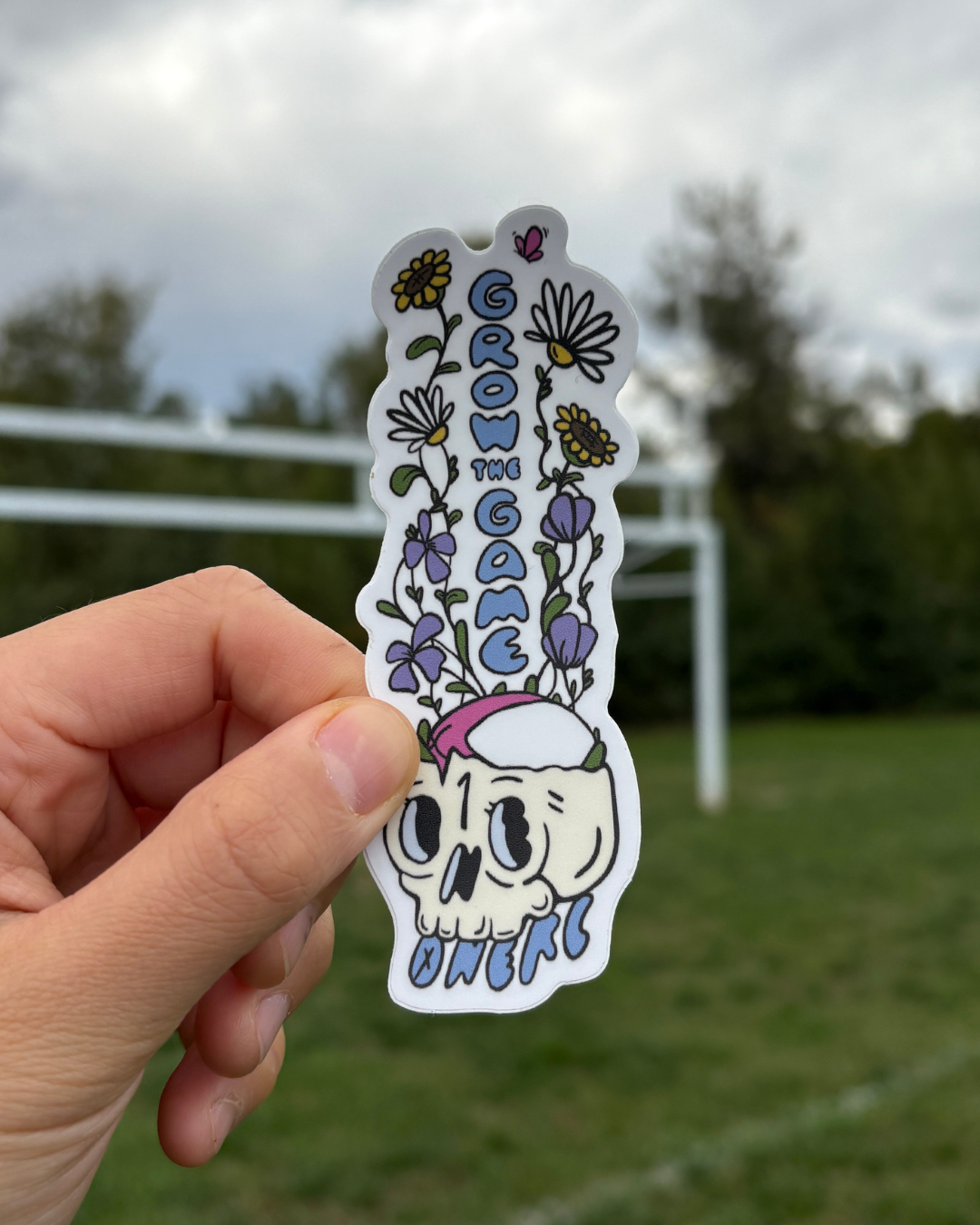 Hand holding a sticker of wildflowers growing out of a skull with a rugby ball resting in it with rugby posts in the background.