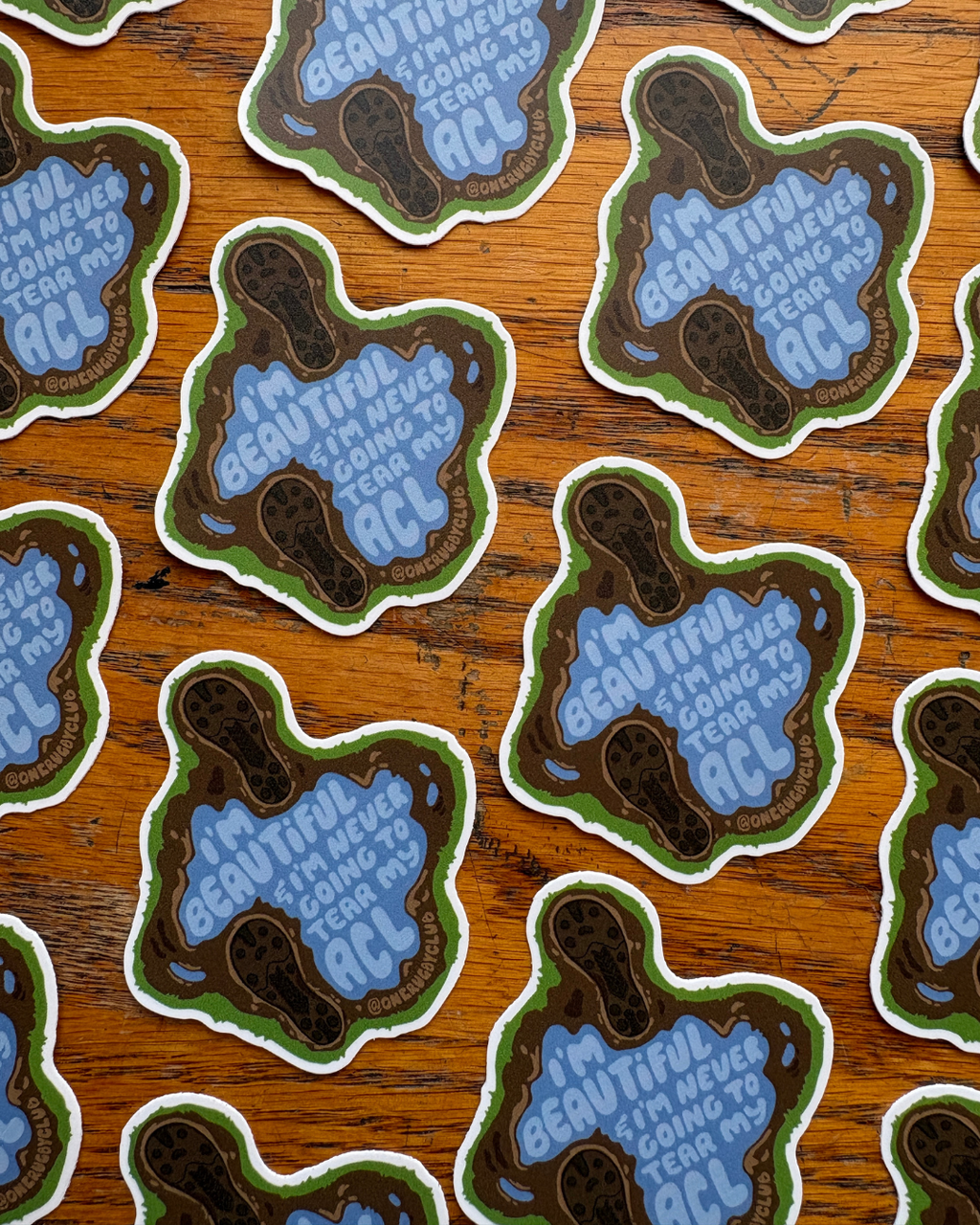 Repeating rows of stickers of muddy cleat prints around a puddle that reads "I'm beautiful & I'm never going to tear my ACL"
