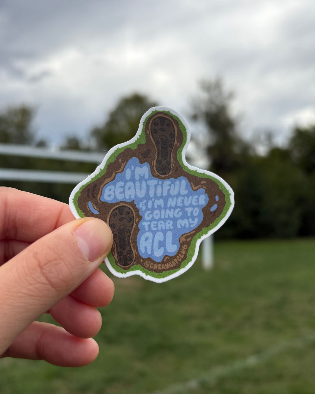 Hand holding sticker of muddy cleat prints around a puddle that reads "I'm beautiful & I'm never going to tear my ACL" with rugby posts in the background