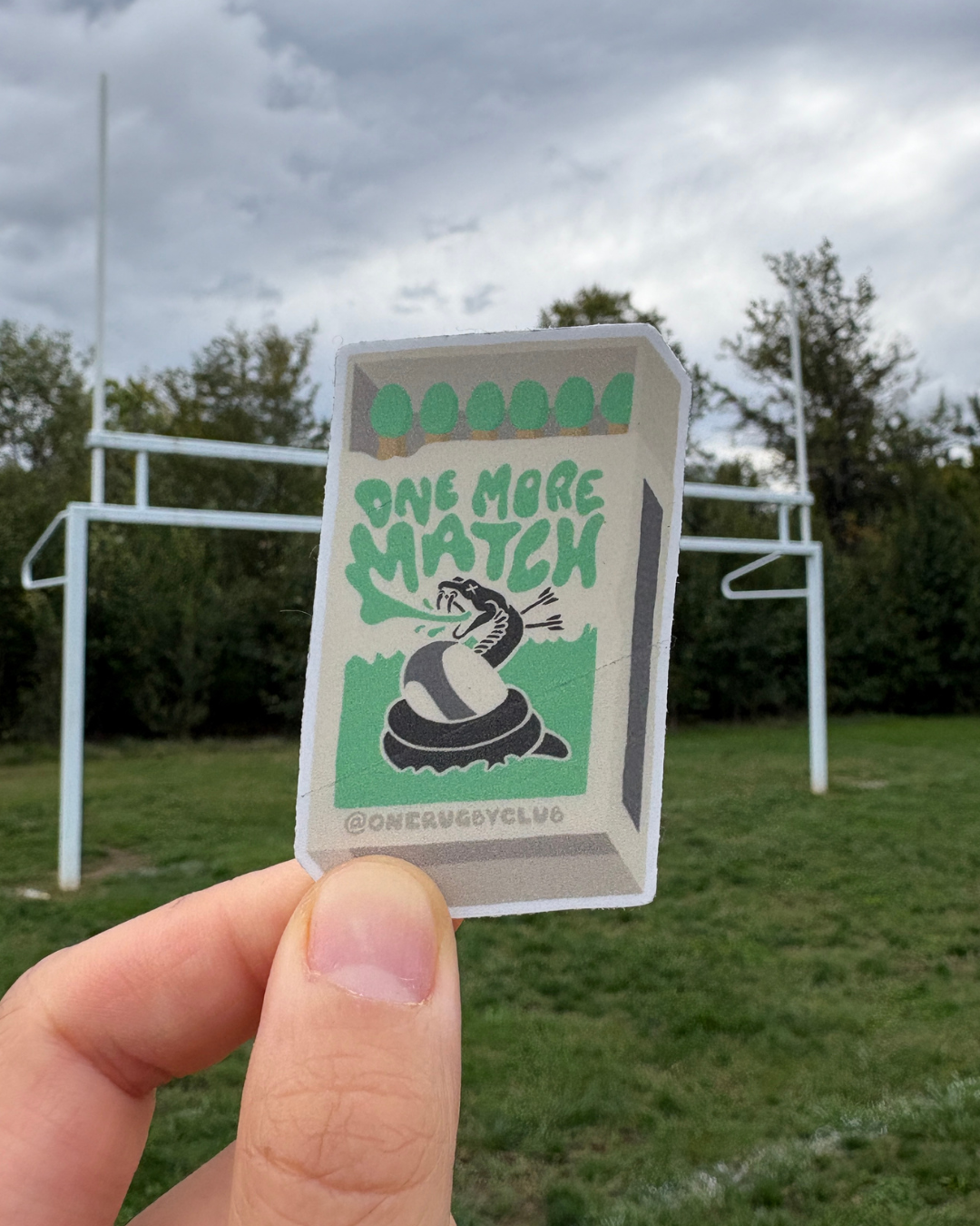 Hand holding sticker of matchbox with venomous snake and the words "One more match" with rugby posts in the background