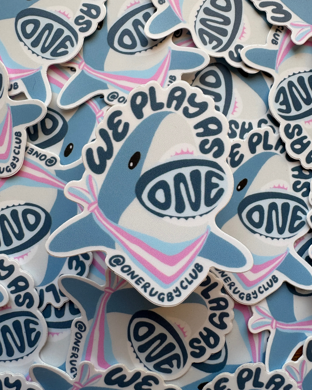 Pile of trans-color, "We Play As One", rugby shark stickers.