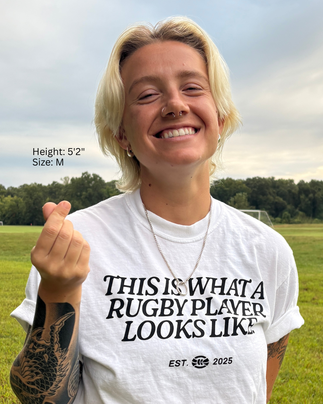 Woman in a grassy field wearing a white t-shirt with the words "This Is What A Rugby Player Looks Like" across the chest.