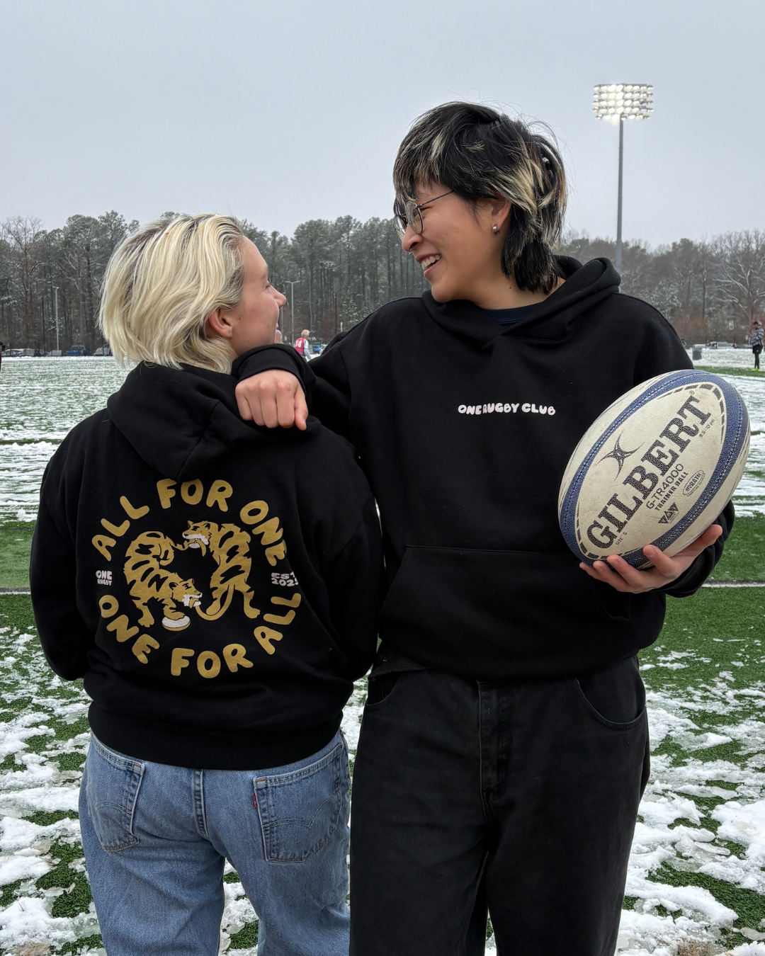 Two women in standing next to each other on a snowy rugby pitch wearing "All for One, One for All" hoodies.