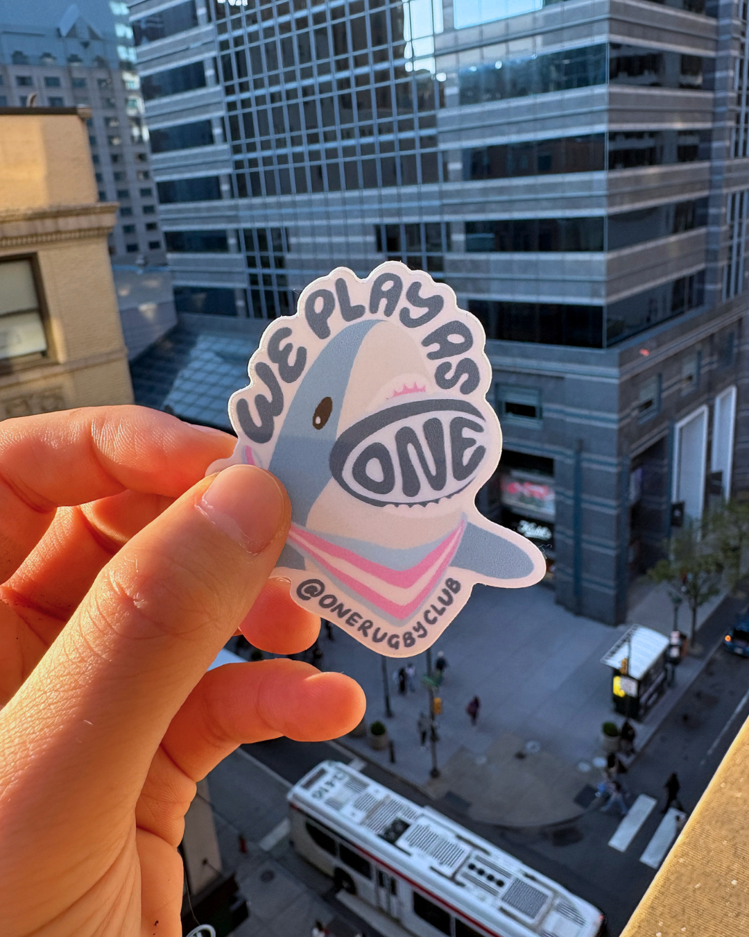 Handing holding trans-color, "We Play As One", rugby shark sticker with a city in the background.