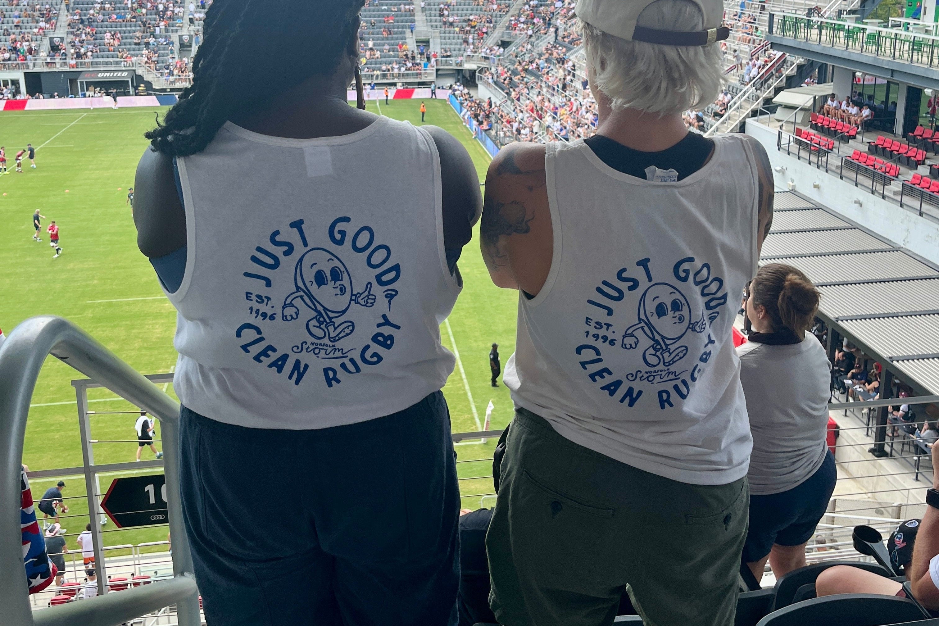 Two women wearing custom "Just Good Clean Rugby" shirts at a USA vs. Fiji women's rugby game.