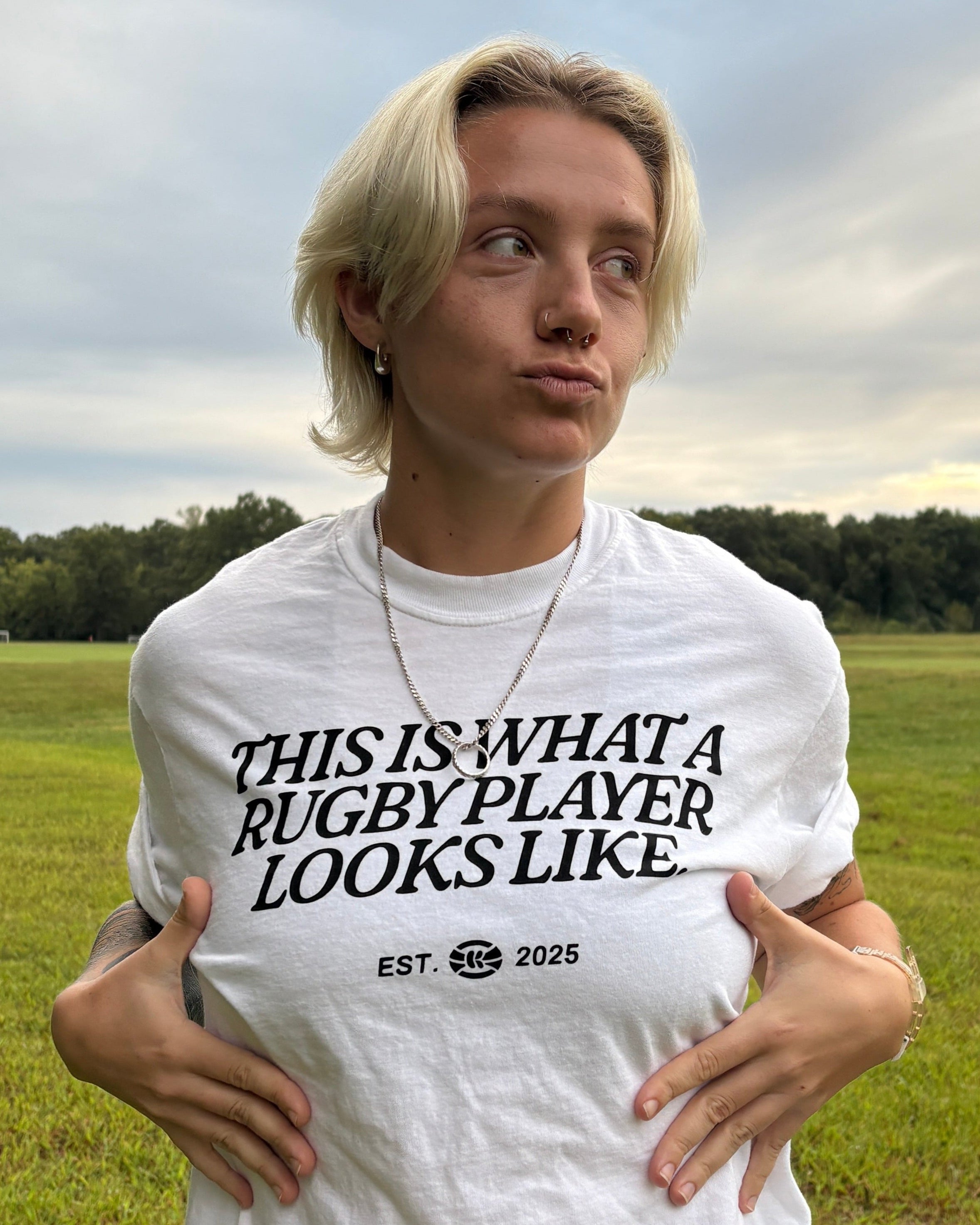 Woman in a grassy field wearing a white t-shirt with the words "This Is What A Rugby Player Looks Like" across the chest.