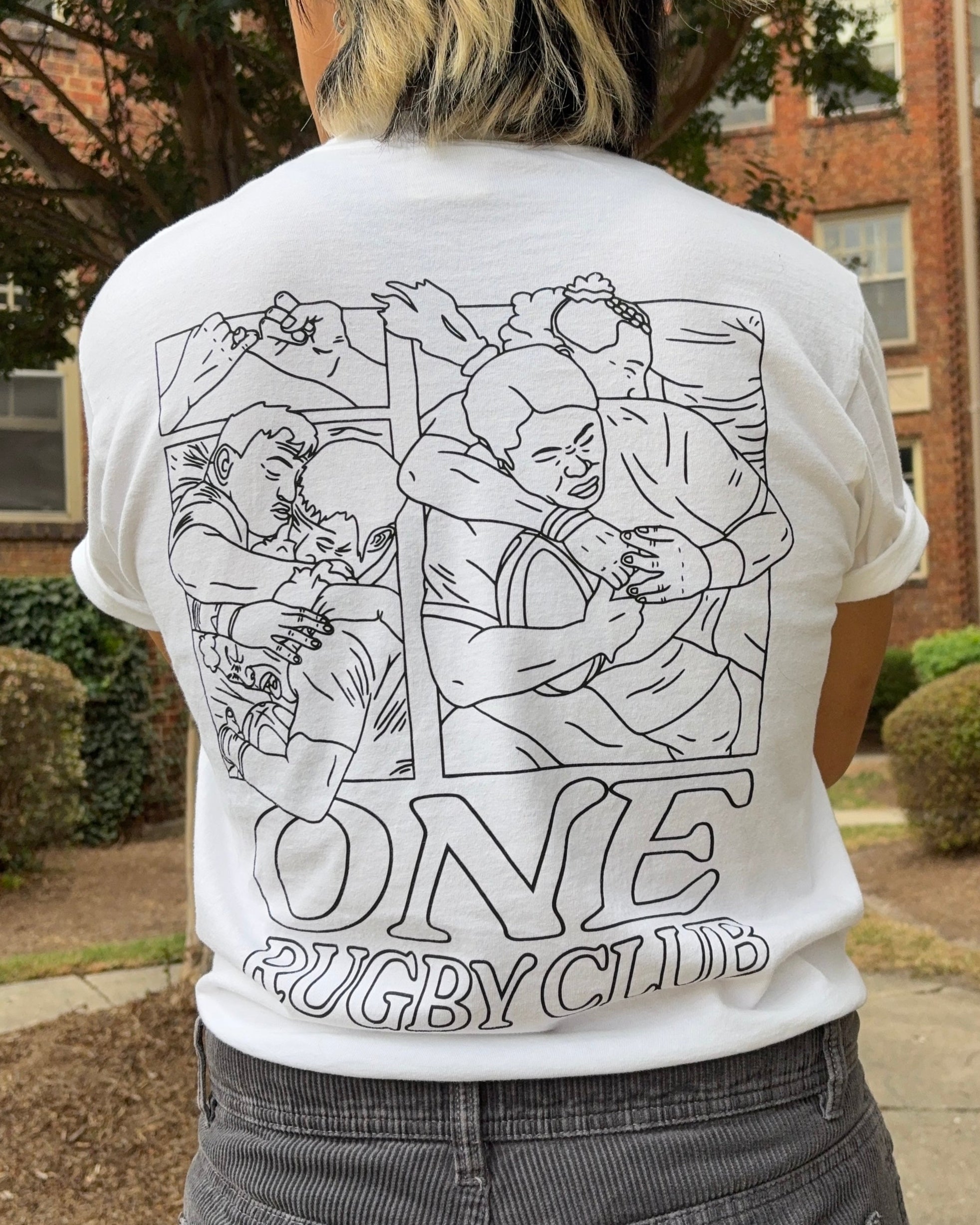 Person with their back facing the camera, wearing a white t-shirt with drawings of rugby players and the words "One Rugby Club" on it.